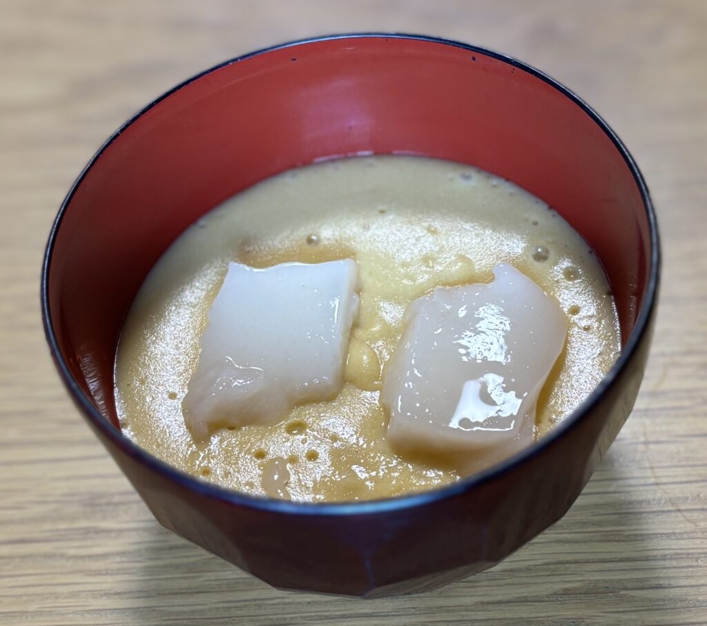 汁椀に盛ったムースのお雑煮の上に、スプーンで食べるおもちを2切れのせた状態。餅は沈まずに浮いている