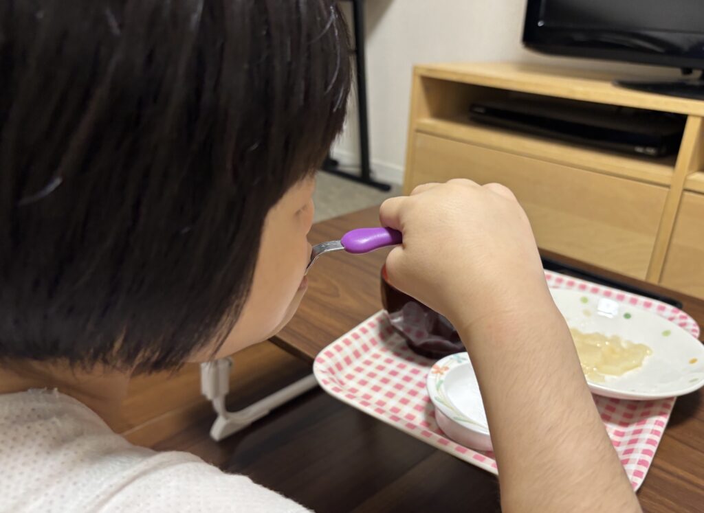 娘が自分でスプーンを持ち、ムースのお雑煮を口に運んでいるところ。手前に汁椀とお餅をのせた小皿がある